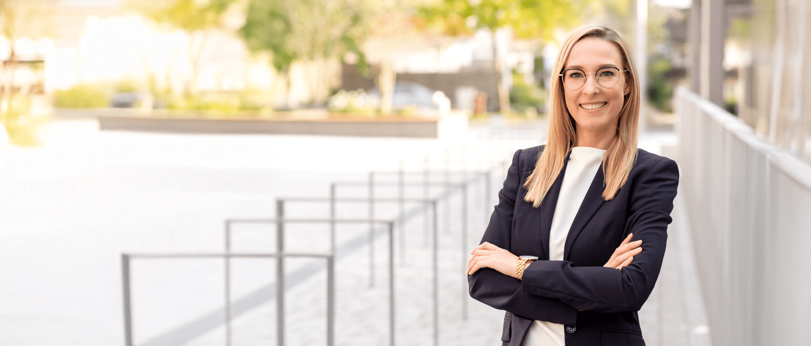 Frau mit langen blonden Haaren und Brille, die in Business-Kleidung vor einem modernen Hintergrund steht.