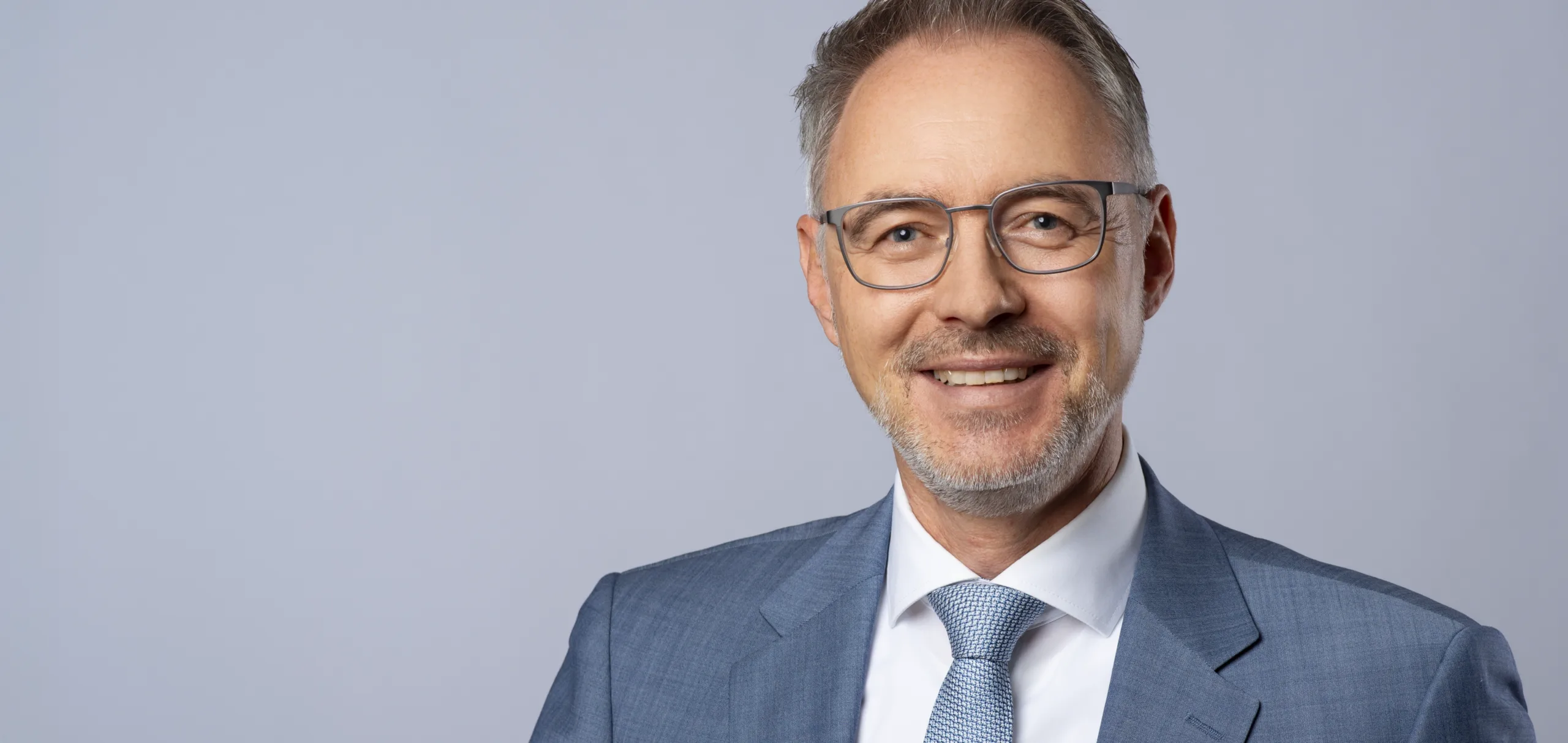 Mann mit Brille und Anzug lächelt in die Kamera vor einem grauen Hintergrund.