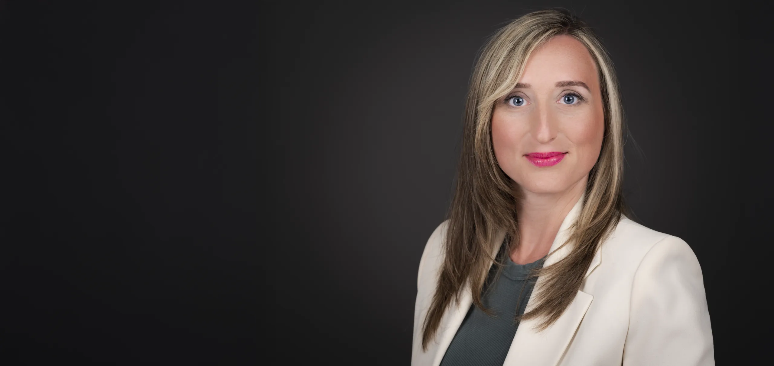 Frau mit langen, blonden Haaren und einem weißen Blazer vor einem dunklen Hintergrund.