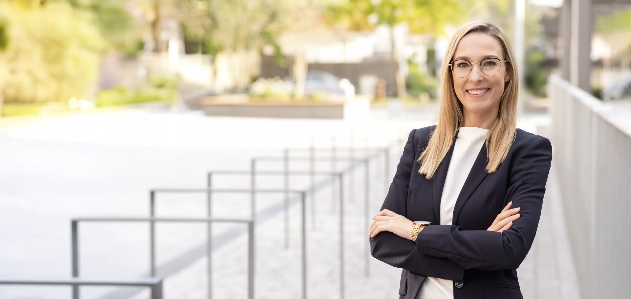 Frau mit Brille und Blazer steht mit verschränkten Armen und lächelt in die Kamera.